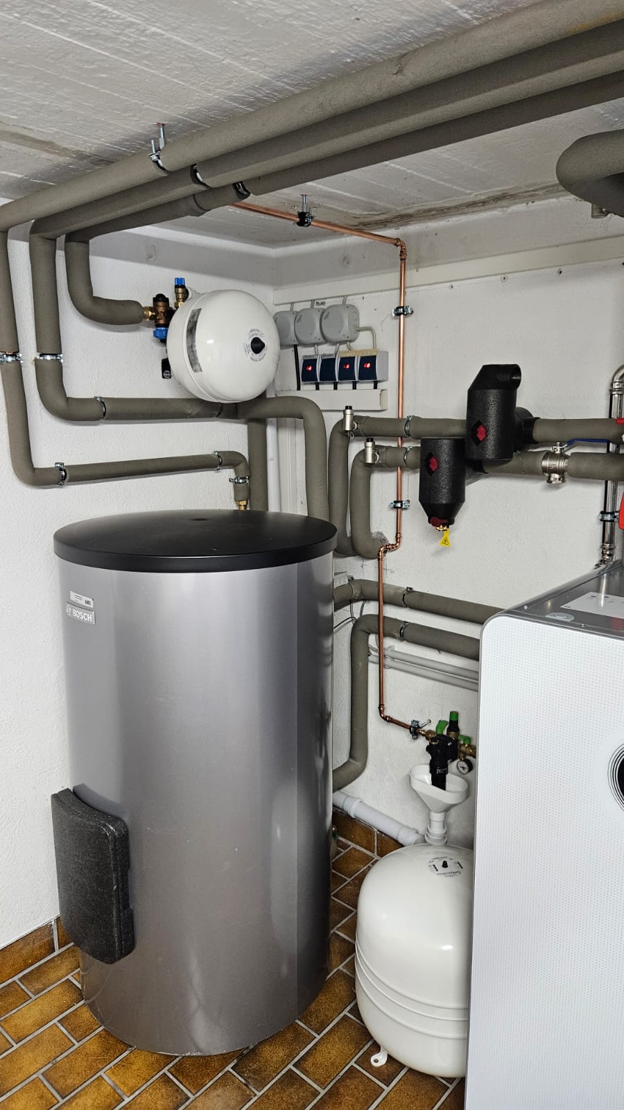 Zu sehen ist eine installierte Bosch-Wärmepumpe mit angeschlossenem Pufferspeicher und sorgfältig gedämmter Rohrleitungstechnik.